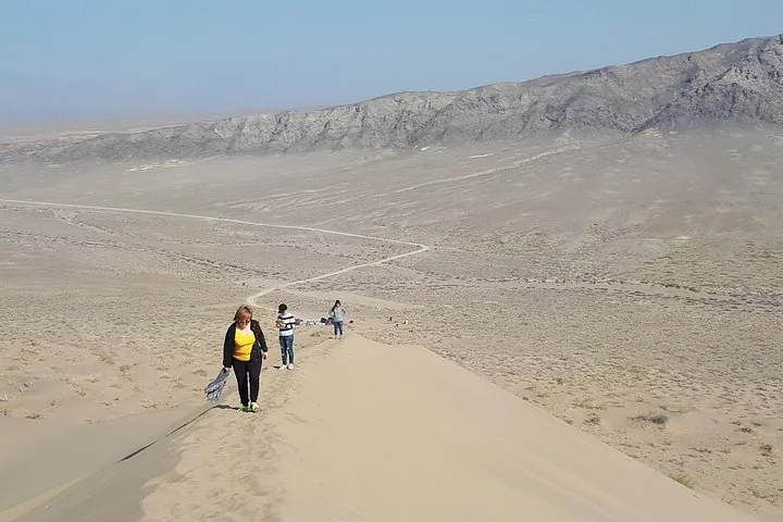 Menschen gehen entlang des Kamms einer Sanddüne, unten erstreckt sich eine weite Ebene.